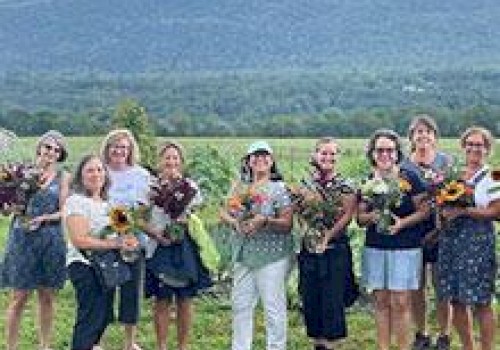 A group of people holding flowers stand in front of a scenic landscape with mountains and greenery under a partly cloudy sky.