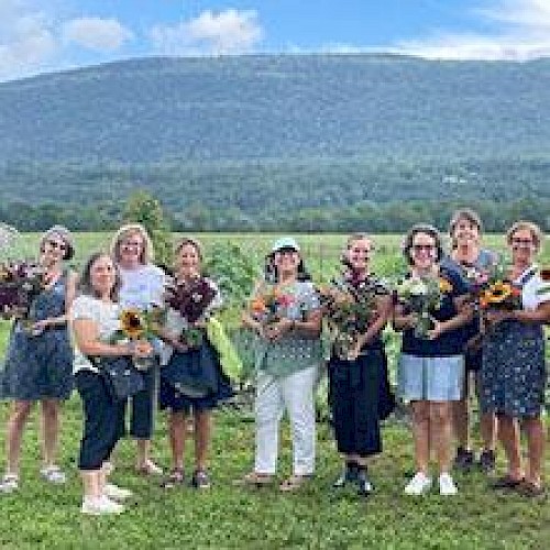 A group of people holding flowers stand in front of a scenic landscape with mountains and greenery under a partly cloudy sky.