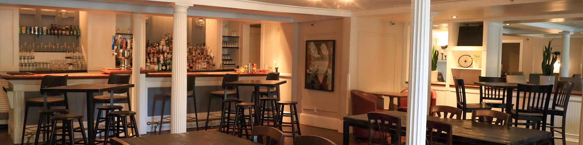 The image shows a spacious restaurant interior with wooden tables, chairs, and a bar area, creating a cozy dining atmosphere.