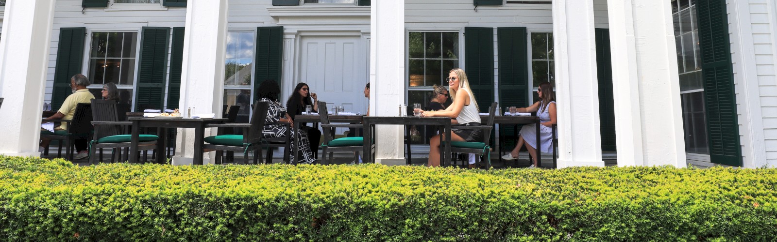 People are dining on a porch of a white building with tall columns and green shutters, surrounded by greenery.