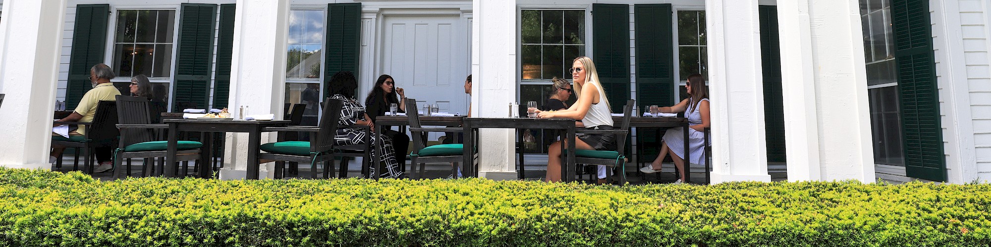 People are dining on a porch of a white building with tall columns and green shutters, surrounded by greenery.