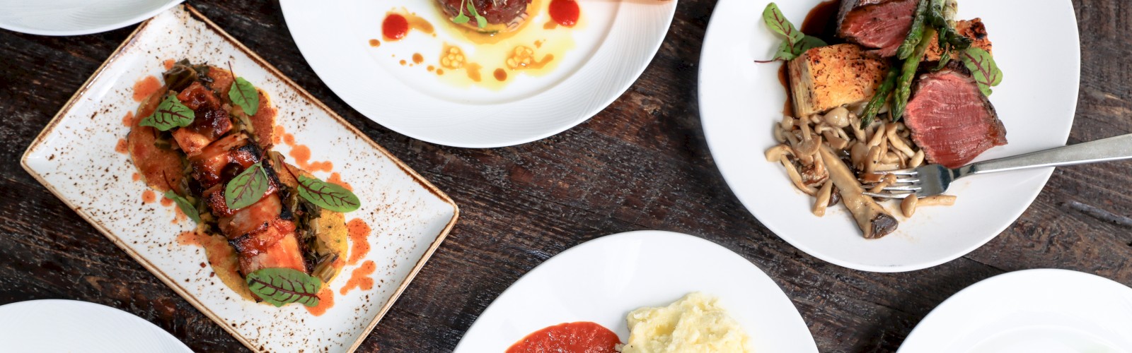 An array of gourmet dishes, including meats, greens, and pasta, presented on white plates atop a wooden surface.