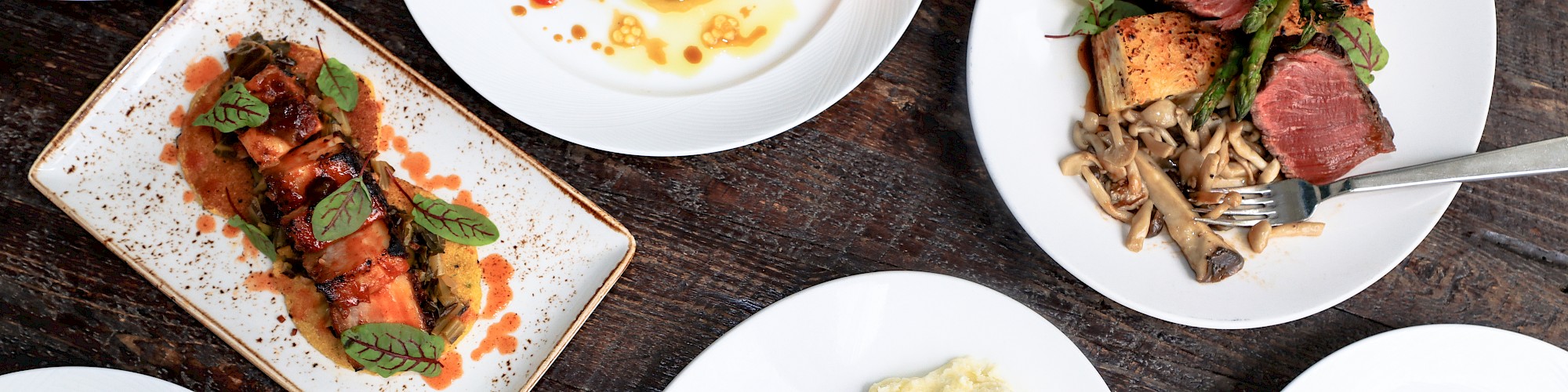 An array of gourmet dishes, including meats, greens, and pasta, presented on white plates atop a wooden surface.