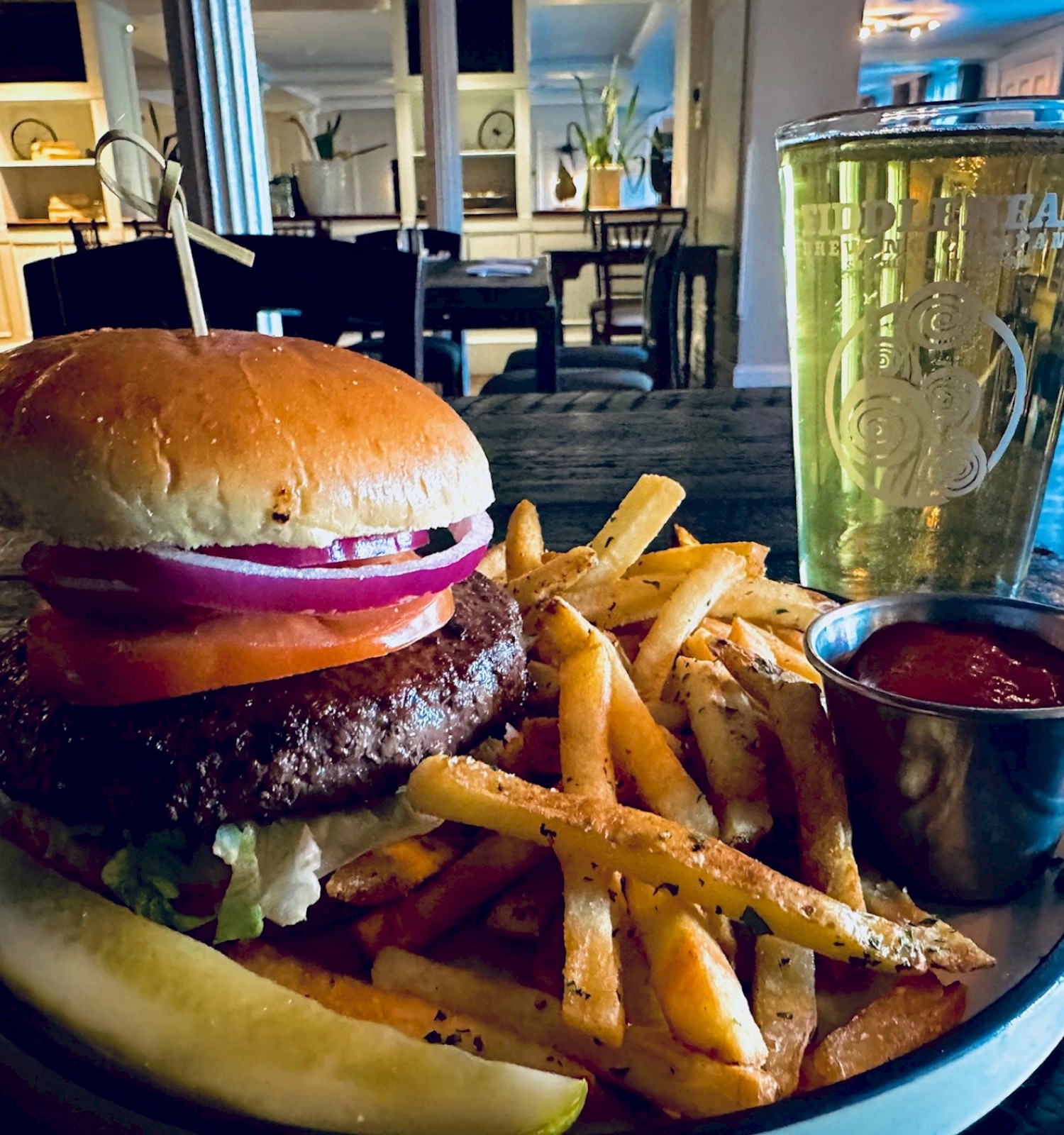 A burger with onion, tomato, and lettuce, accompanied by fries, a pickle, ketchup, and a glass of beer in a cozy setting.