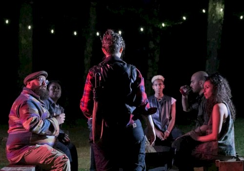 A group of people are gathered outdoors at night, sitting in a circle under string lights, with one person standing in the center speaking.