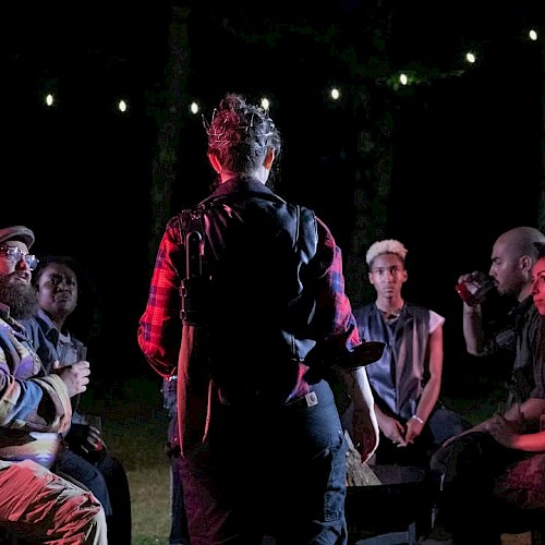 A group of people are gathered outdoors at night, sitting in a circle under string lights, with one person standing in the center speaking.