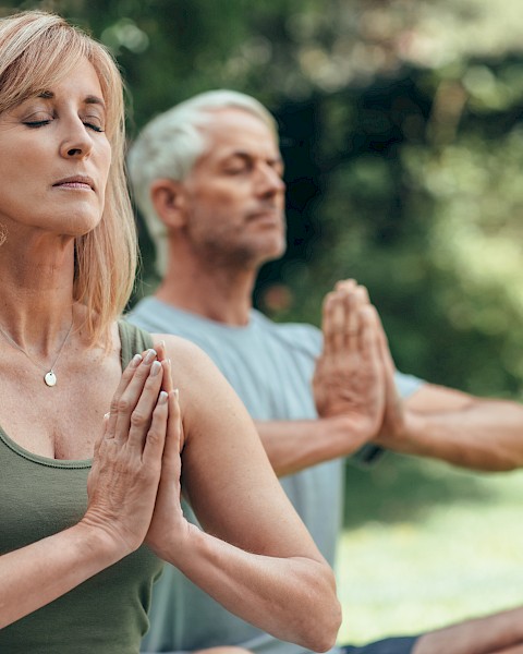 Two people are practicing yoga outdoors with hands in prayer position, eyes closed, and a peaceful expression.