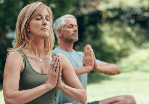 Two people are practicing yoga outdoors with hands in prayer position, eyes closed, and a peaceful expression.