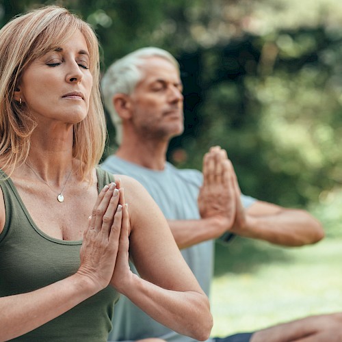 Two people are practicing yoga outdoors with hands in prayer position, eyes closed, and a peaceful expression.