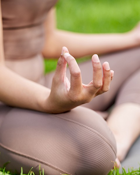 A person is practicing yoga outdoors, sitting cross-legged on a mat with hands in a mudra, surrounded by grass.
