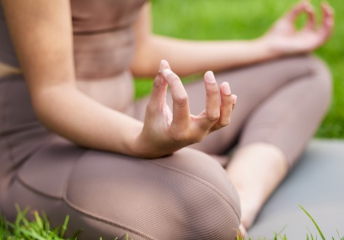 A person is practicing yoga outdoors, sitting cross-legged on a mat with hands in a mudra, surrounded by grass.