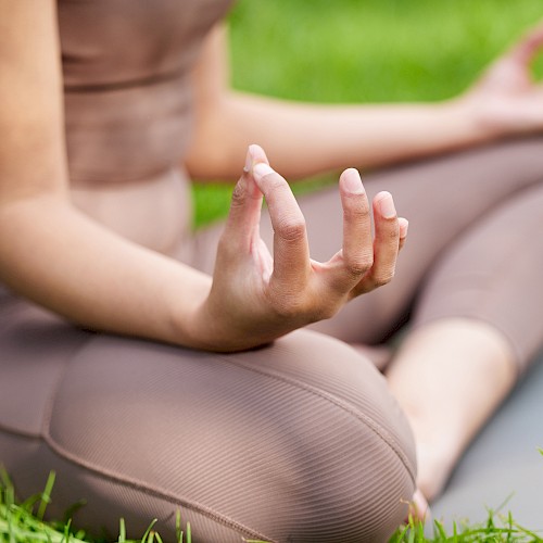 A person is practicing yoga outdoors, sitting cross-legged on a mat with hands in a mudra, surrounded by grass.