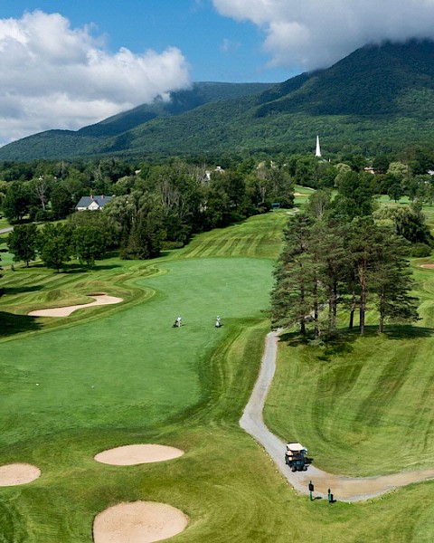 A scenic golf course with well-maintained greens, sand bunkers, and a golf cart on a path. Mountains in the background under a partly cloudy sky.