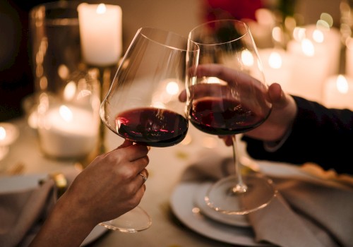 Two people clinking glasses of red wine at a candlelit table, surrounded by cozy, warm lighting and an elegant dinner setting.