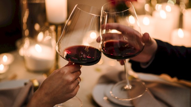 Two people clinking glasses of red wine at a candlelit table, surrounded by cozy, warm lighting and an elegant dinner setting.