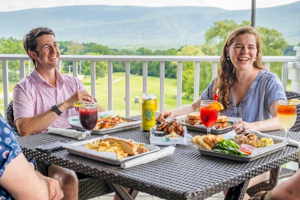 Four people are enjoying a meal outdoors at a table with drinks and food, overlooking a scenic landscape with mountains in the background.