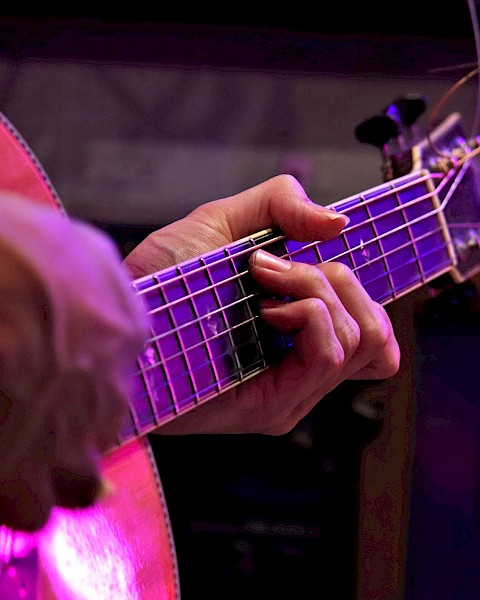 A person is playing an acoustic guitar, captured in a close-up with a focus on their hands and strings, under purple lighting.