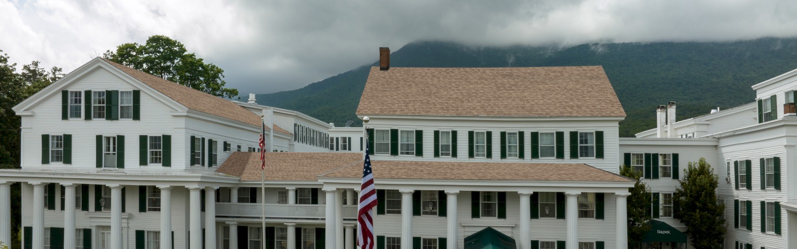 A historic building with white columns and antique cars in front, set against a backdrop of clouds and mountains.