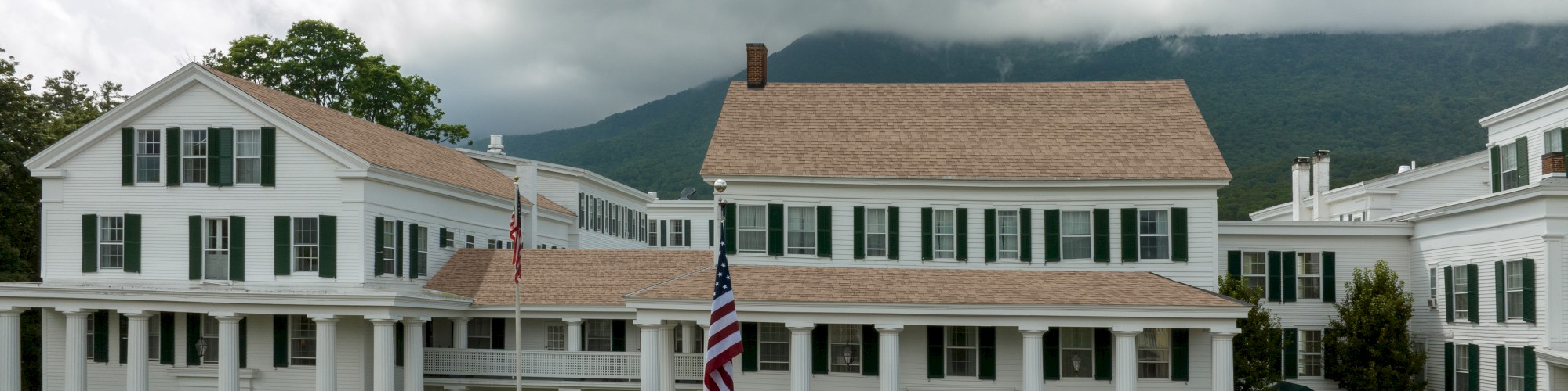 A historic building with white columns and antique cars in front, set against a backdrop of clouds and mountains.