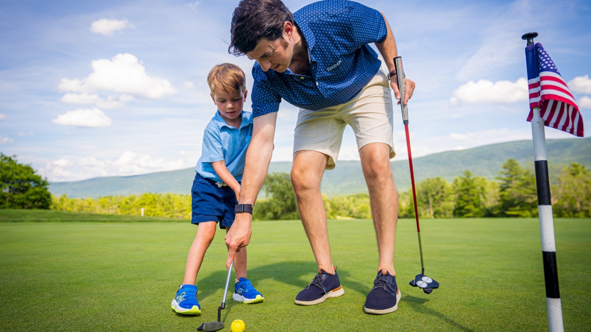 A man is helping a child putt a golf ball on a green. They're near a hole with a flag, under a blue sky with scattered clouds.