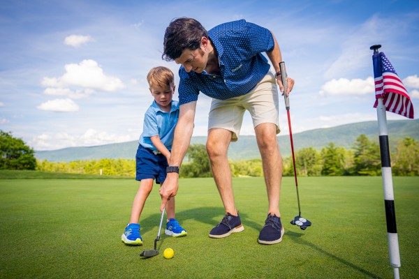 An adult and child are on a golf course putting a ball near a hole with a flag. The scene is sunny with a backdrop of trees and hills.