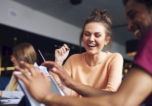 A group of people are smiling and interacting around a tablet in a casual setting, suggesting collaboration or a friendly meeting.