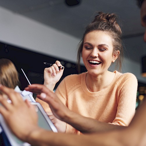A group of people are smiling and interacting around a tablet in a casual setting, suggesting collaboration or a friendly meeting.
