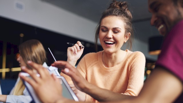 A group of people are smiling and interacting around a tablet in a casual setting, suggesting collaboration or a friendly meeting.