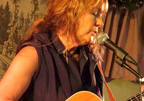 A person with reddish hair is playing an acoustic guitar and singing into a microphone, with a decorative background behind them.