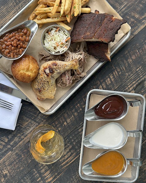 A tray with ribs, fries, coleslaw, chicken, beans, and cornbread; sauces and a drink are on the side.