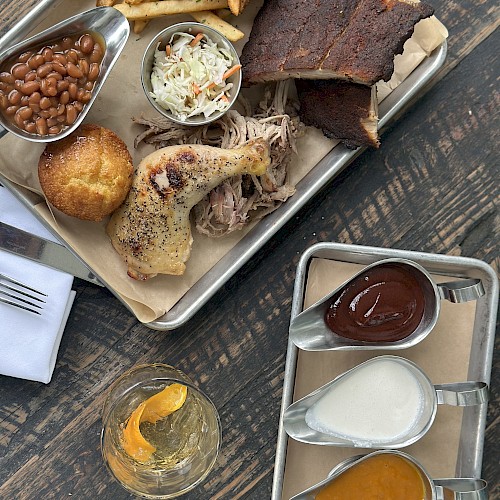 A tray with ribs, fries, coleslaw, chicken, beans, and cornbread; sauces and a drink are on the side.