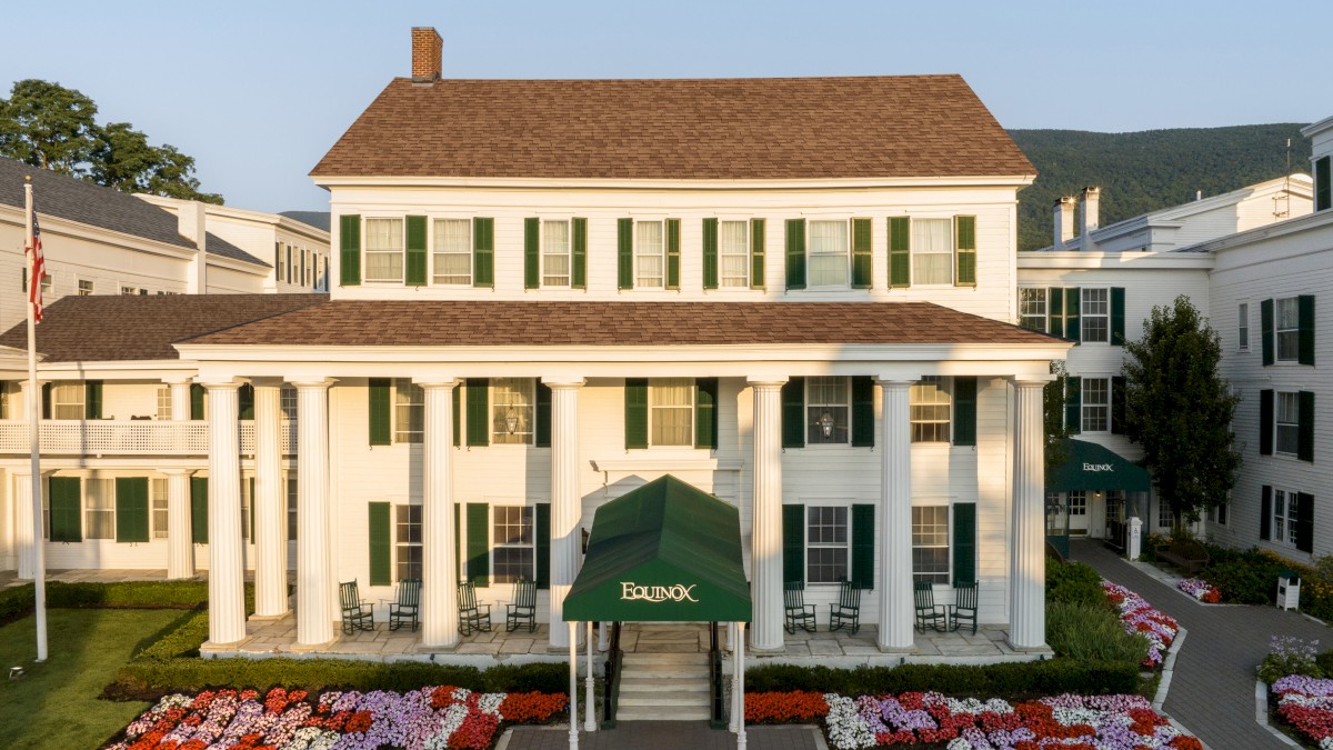A large white building with green shutters and an entrance canopy. Surrounding it are bright flower beds, with a clear sky in the background.
