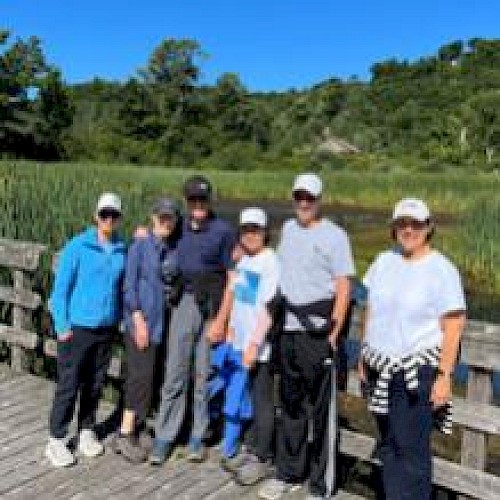 A group of six people wearing casual outdoor clothing stands on a wooden deck with greenery in the background.
