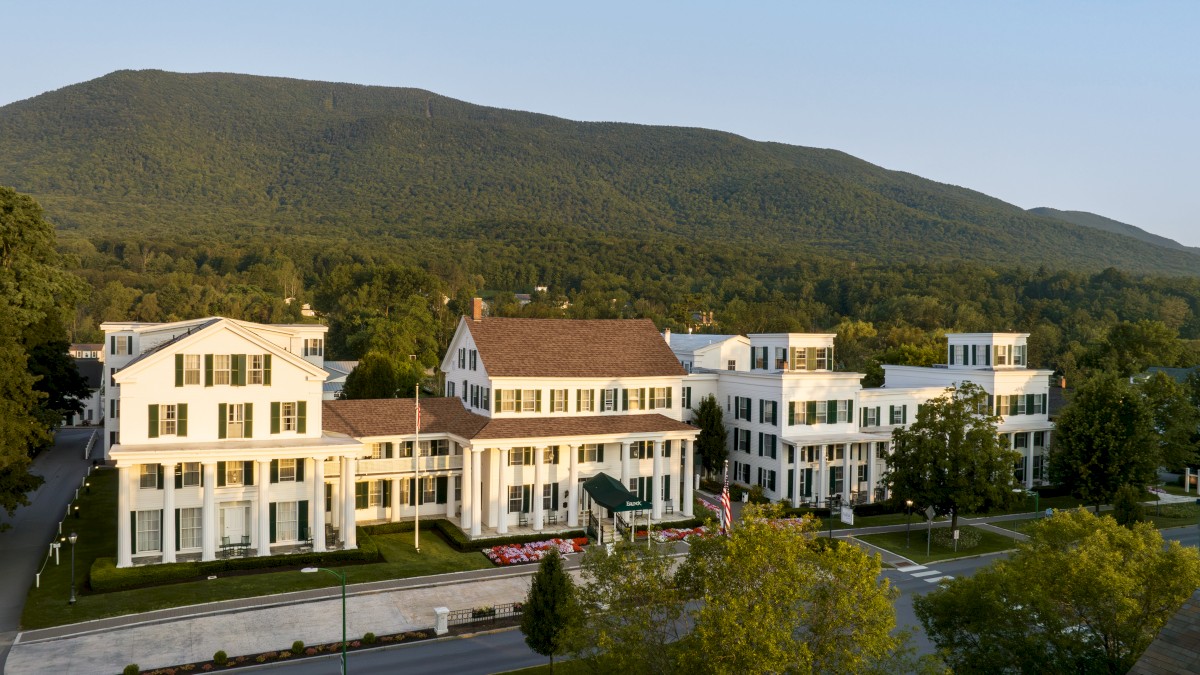 A large white building with columns sits amidst lush greenery, with a mountain in the background and a clear sky above.