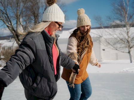 Two people in winter clothing are skating on ice outside, with trees and a building in the snowy background.