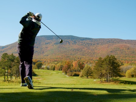 A person swings a golf club on a green golf course with scenic autumnal trees and mountains in the background under a clear blue sky.