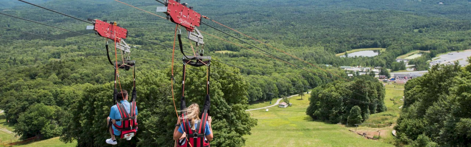 People are riding zipline chairs over a green landscape with mountains in the background. The scene is set outdoors.