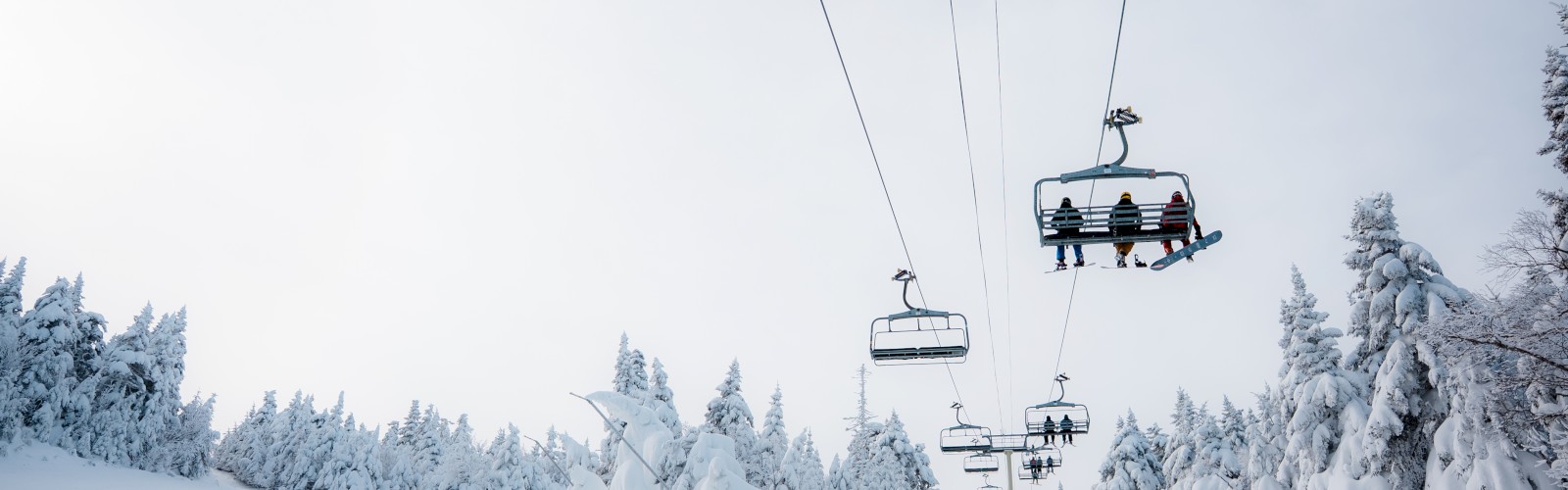 A snowy landscape with a ski lift carrying people over a slope, surrounded by snow-covered trees under a cloudy sky.