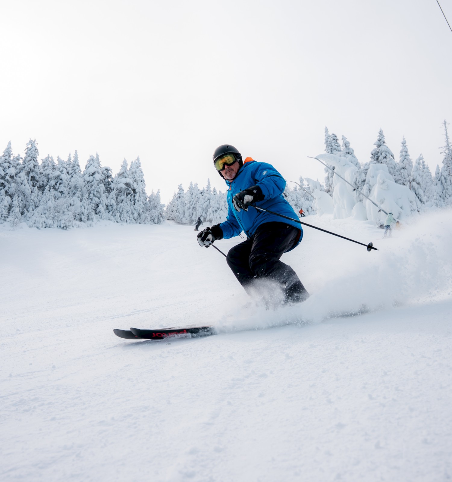 A person is skiing downhill on a snowy slope with trees in the background, wearing winter gear and goggles, with ski lifts overhead.