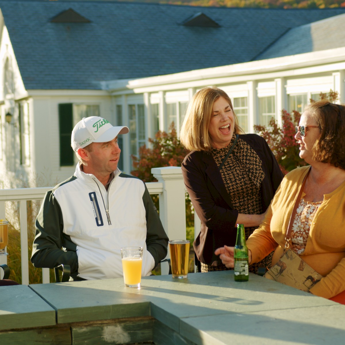Four people are sitting outside, enjoying drinks and talking, with a building and autumn trees in the background.