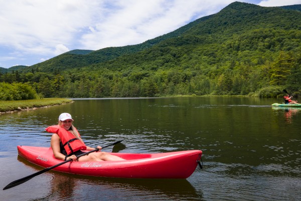 A person kayaking on a calm lake, surrounded by lush green hills and a partly cloudy sky.