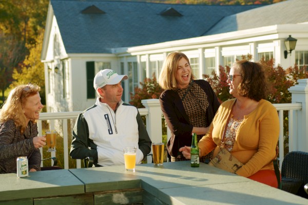 A group of four people enjoying drinks and chatting outside a building with autumn foliage in the background.