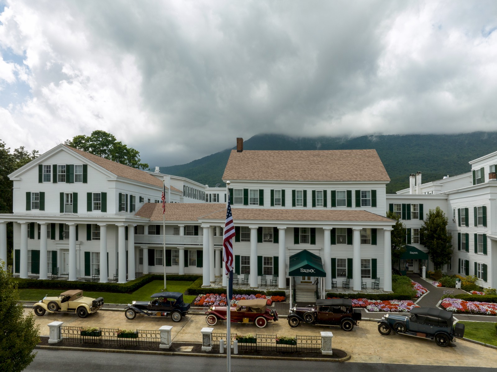 A classic hotel with vintage cars parked in front, surrounded by dramatic clouds and mountains in the background.