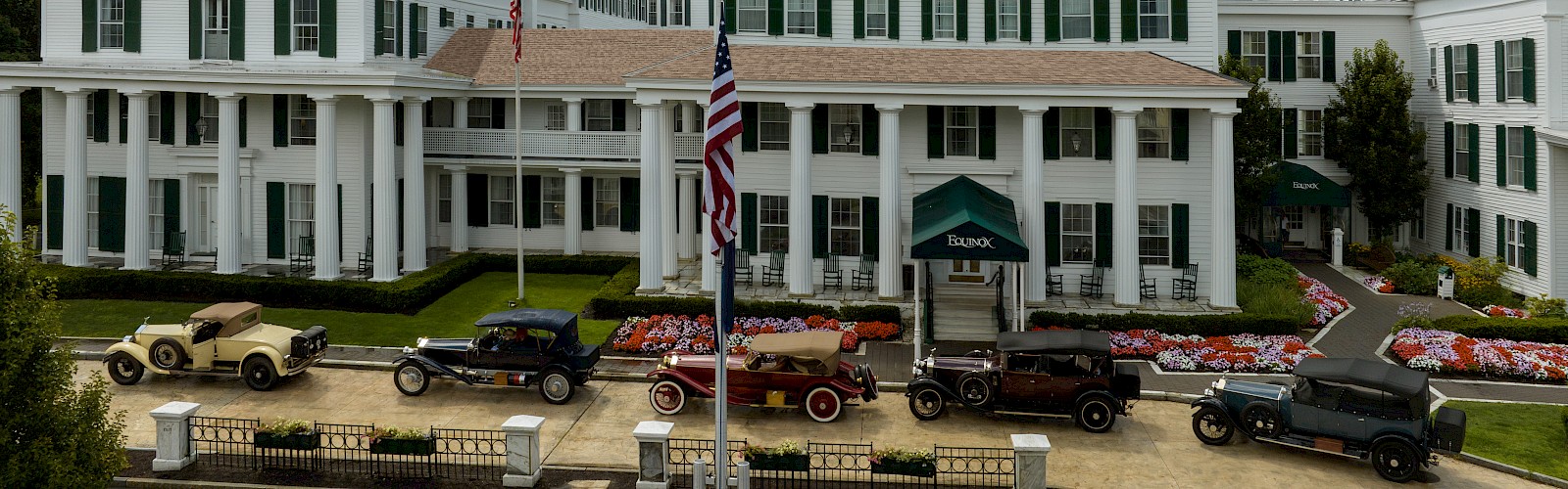 A classic hotel with vintage cars parked in front, surrounded by dramatic clouds and mountains in the background.