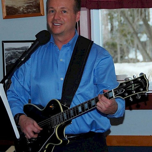 A person in a blue shirt is playing an electric guitar and singing into a microphone in a room with framed pictures on the wall.