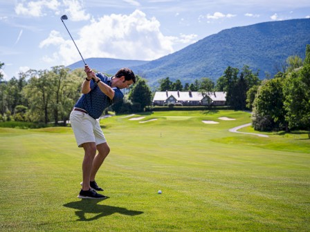 A person is swinging a golf club on a lush golf course with mountains in the background under a partly cloudy sky.