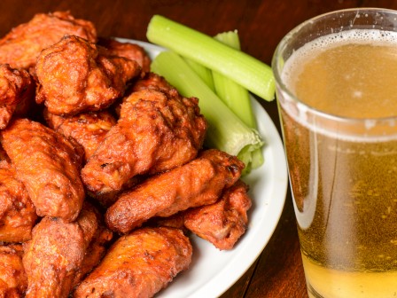 A plate of buffalo wings with celery sticks next to a glass of beer on a wooden surface.