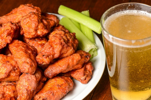 A plate of buffalo wings with celery sticks next to a glass of beer on a wooden surface.