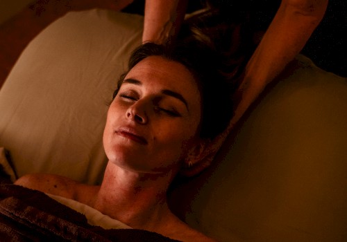 A person is receiving a relaxing massage in dim lighting while lying comfortably with their eyes closed under a brown blanket.