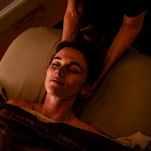 A person is receiving a relaxing massage in dim lighting while lying comfortably with their eyes closed under a brown blanket.